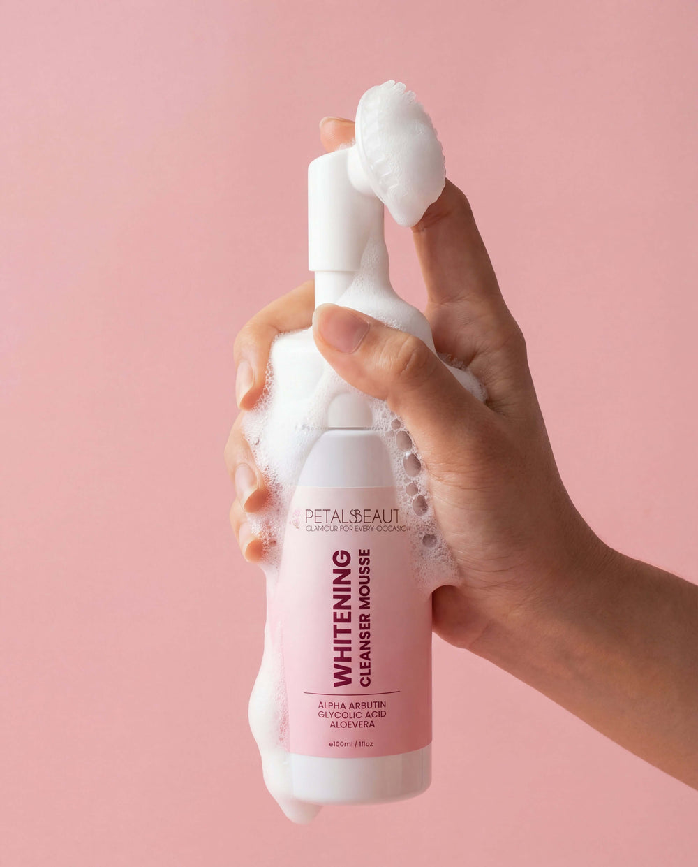 Hand holding a foamy bottle of Petals Beauty Whitening Cleanser Mousse against a pink background
