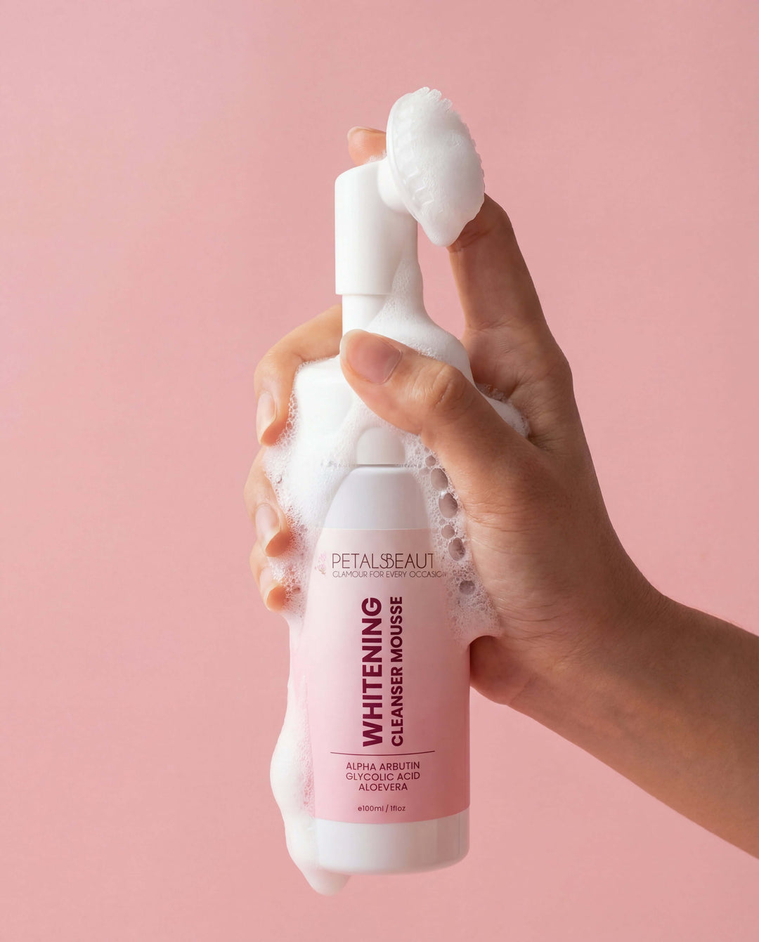 Hand holding a foamy bottle of Petals Beauty Whitening Cleanser Mousse against a pink background